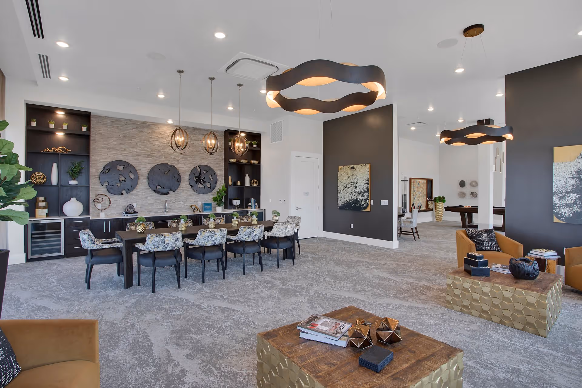 Bright modern communal lounge and dining area with a long table, patterned chairs, pendant lights, and casual seating with coffee tables.