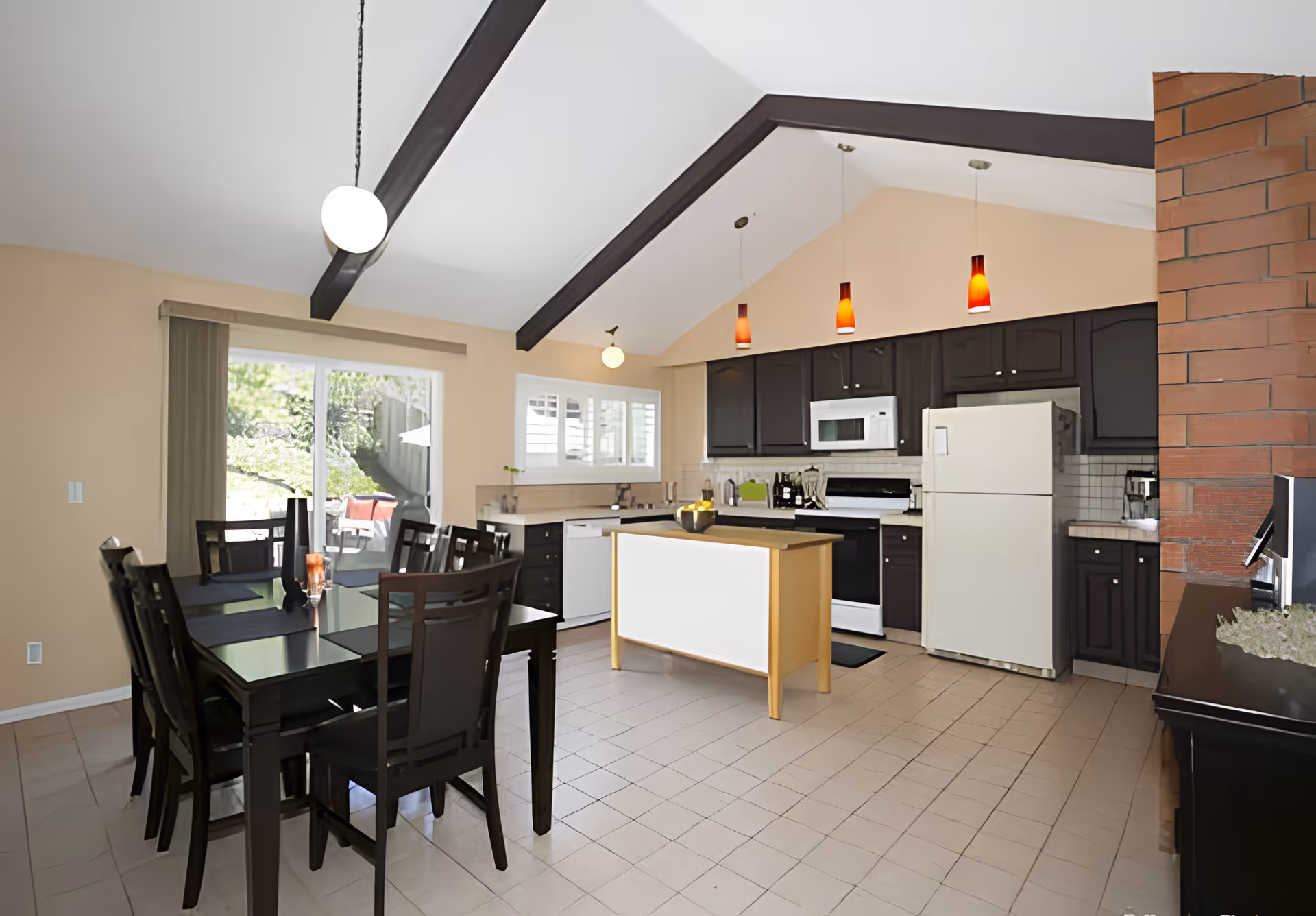 Open-plan kitchen and dining area with a black dining table, central island, dark cabinetry, white appliances and a vaulted ceiling with exposed beams.