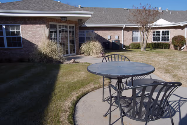 Outdoor patio area at Wheeler Terrace with a round metal table and three matching chairs on a concrete slab. The patio is surrounded by grass and landscaping, with a brick building featuring multiple windows and a glass door in the background under a clear blue sky.