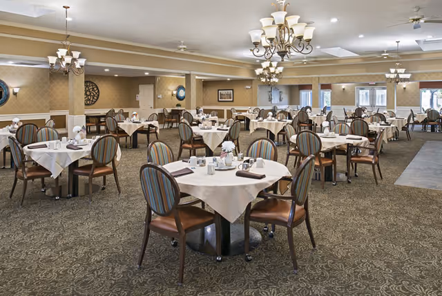 Spacious dining room with multiple round tables covered with white tablecloths, each set with napkins, cups, and condiments. The room features patterned carpet, beige walls with white trim, several chandeliers, and large windows allowing natural light.