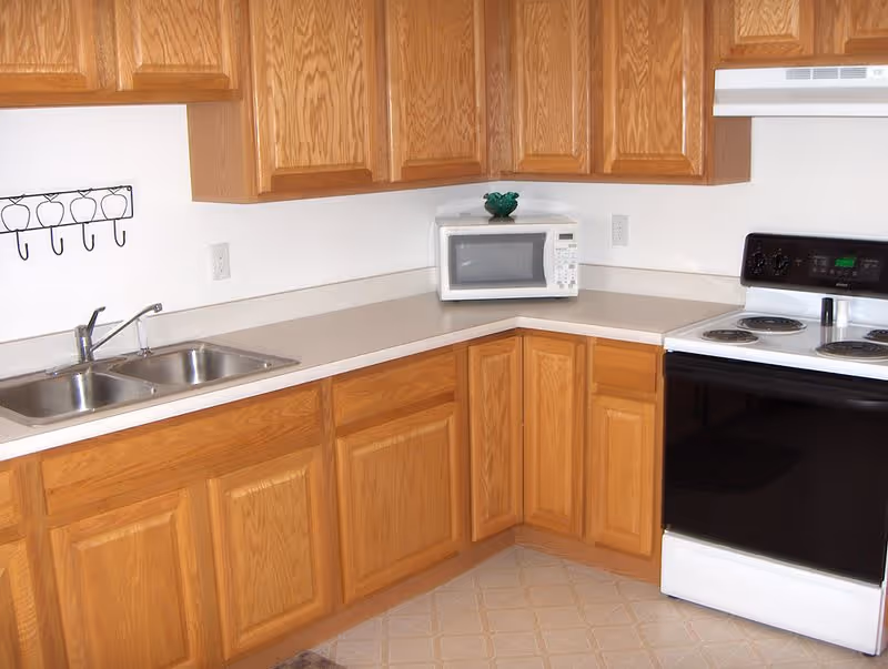 A kitchen corner with wooden cabinets, a double stainless steel sink, a white microwave on the countertop, and a white electric stove with black oven door and control panel.