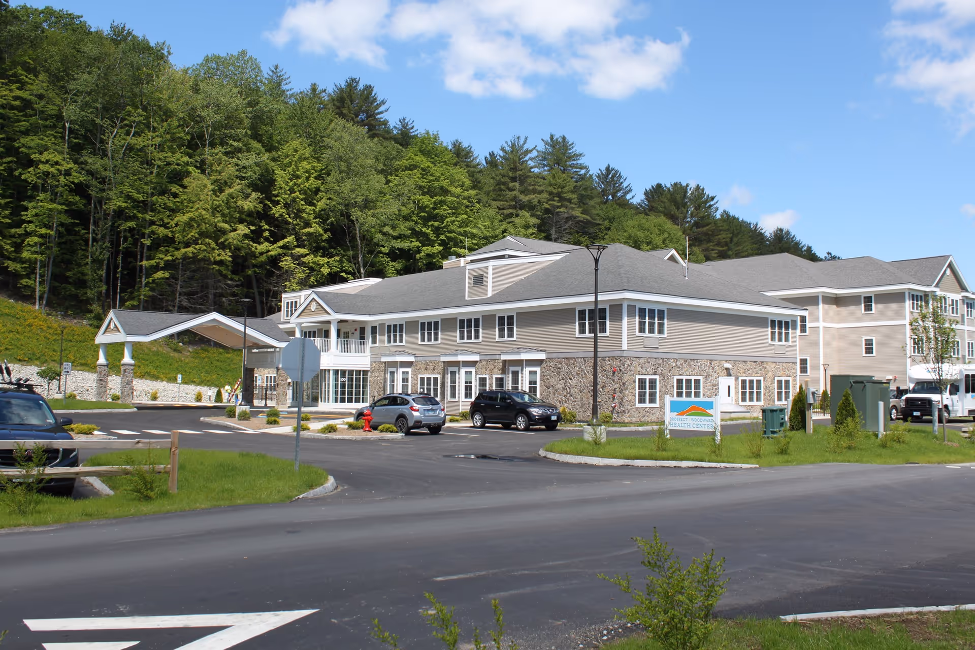 Front exterior of a two-story health center building with parked cars, a driveway, and a wooded hillside behind.
