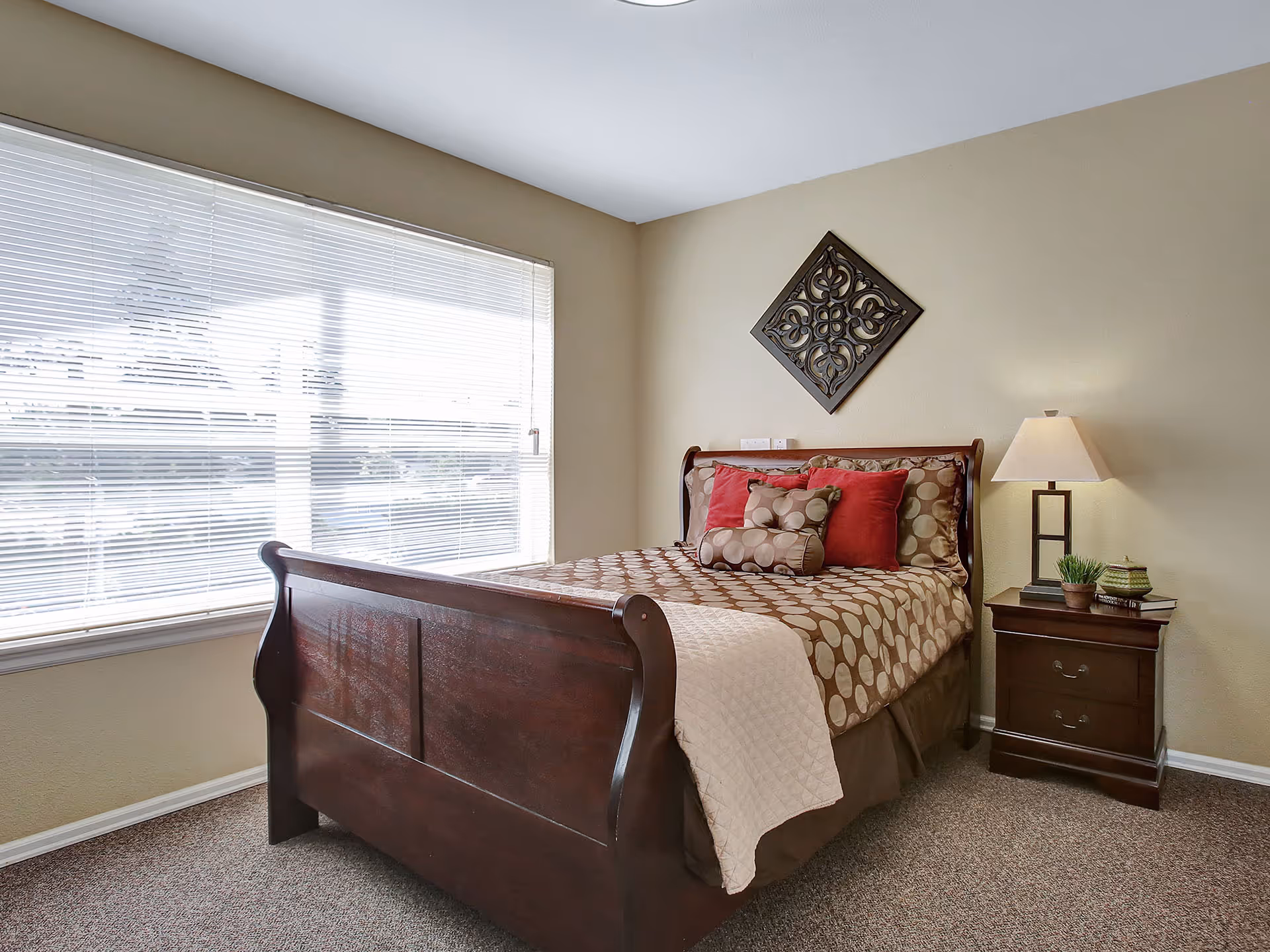 A bedroom with a wooden sleigh bed featuring brown and beige polka dot bedding and red accent pillows. There is a nightstand with a lamp, a small plant, and books next to the bed. A decorative wall piece hangs above the bed, and a large window with blinds lets in natural light.