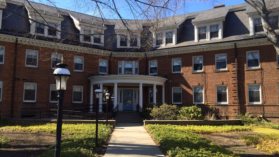 Brick three-story building with a curved portico entrance, steps and lamp posts along a walkway surrounded by landscaping.