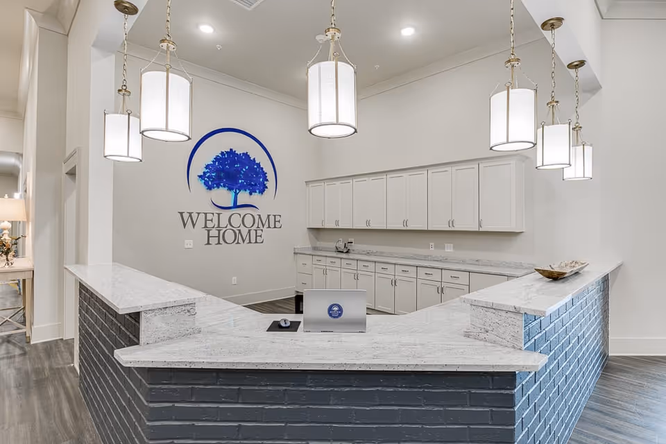 Reception area with a large U-shaped counter featuring a gray brick base and white marble countertop. Above the counter hang six modern pendant lights with white shades. On the wall behind the counter is a blue tree logo with the words 'WELCOME HOME'. White cabinets line the back wall, and a laptop is open on the counter.