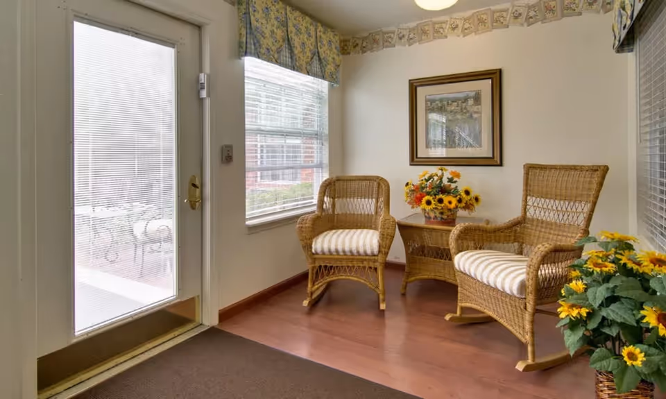 Small sunlit seating area with two wicker chairs, a side table holding a vase of sunflowers, and a glass door to the outside.