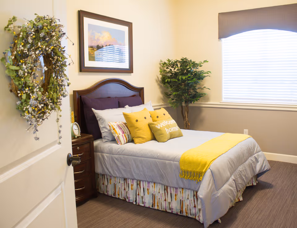 A cozy bedroom with a neatly made bed featuring gray bedding, yellow accent pillows including one that says 'welcome home', and a yellow throw blanket. There is a wooden headboard, a nightstand with a clock, a framed landscape painting on the wall, a green potted plant in the corner, and a window with blinds and a valance. A decorative wreath hangs on the partially open door.