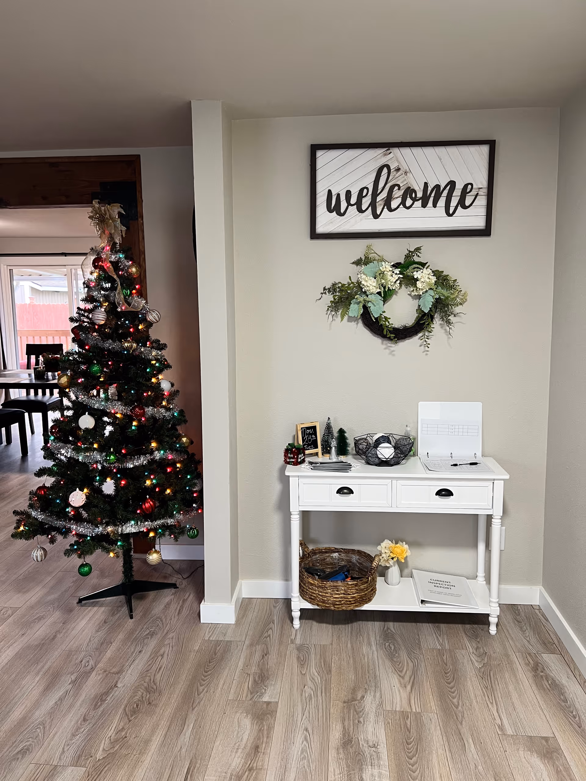 A decorated entry/living area with a lit Christmas tree on the left and a white console table beneath a framed 'welcome' sign and wreath.