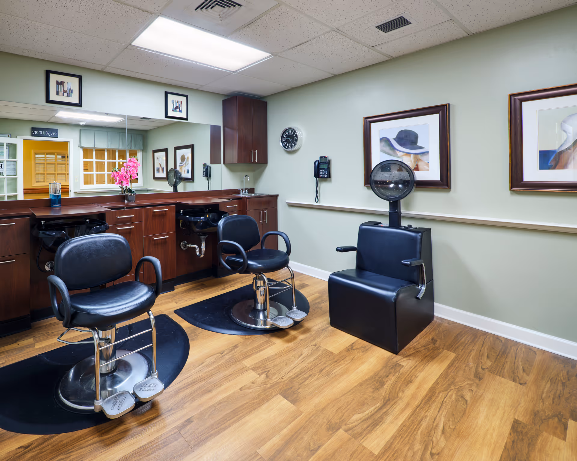Interior view of a salon area in an assisted living facility featuring two black salon chairs in front of a counter with sinks, a hair dryer chair, framed artwork on light green walls, a wall clock, and a telephone.