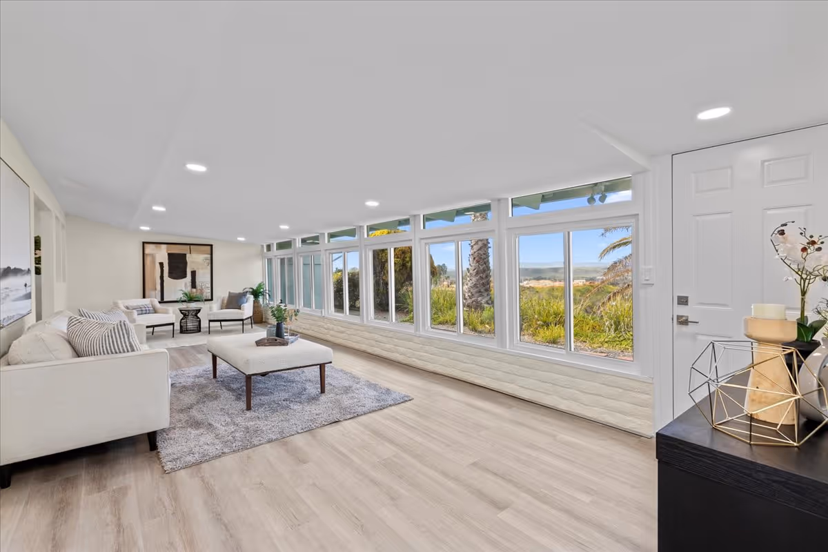 Bright modern living room with sofas, a coffee table, and a long wall of windows overlooking palm trees and a coastal landscape.