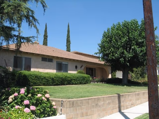 Genesis Manor exterior: a single-story beige building with a manicured lawn, hedges, and trees.