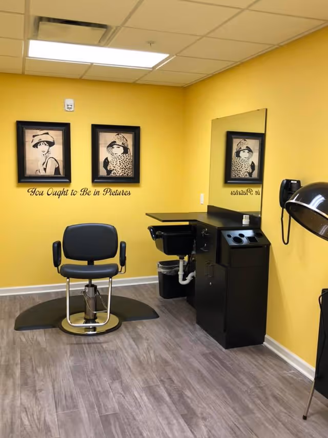 A small hair salon area with a black salon chair on a black mat, a black counter with a sink, a large mirror, and a wall-mounted hair dryer. The walls are painted yellow and decorated with two framed pictures of women wearing hats and the phrase 'You Ought to Be in Pictures' written below them.