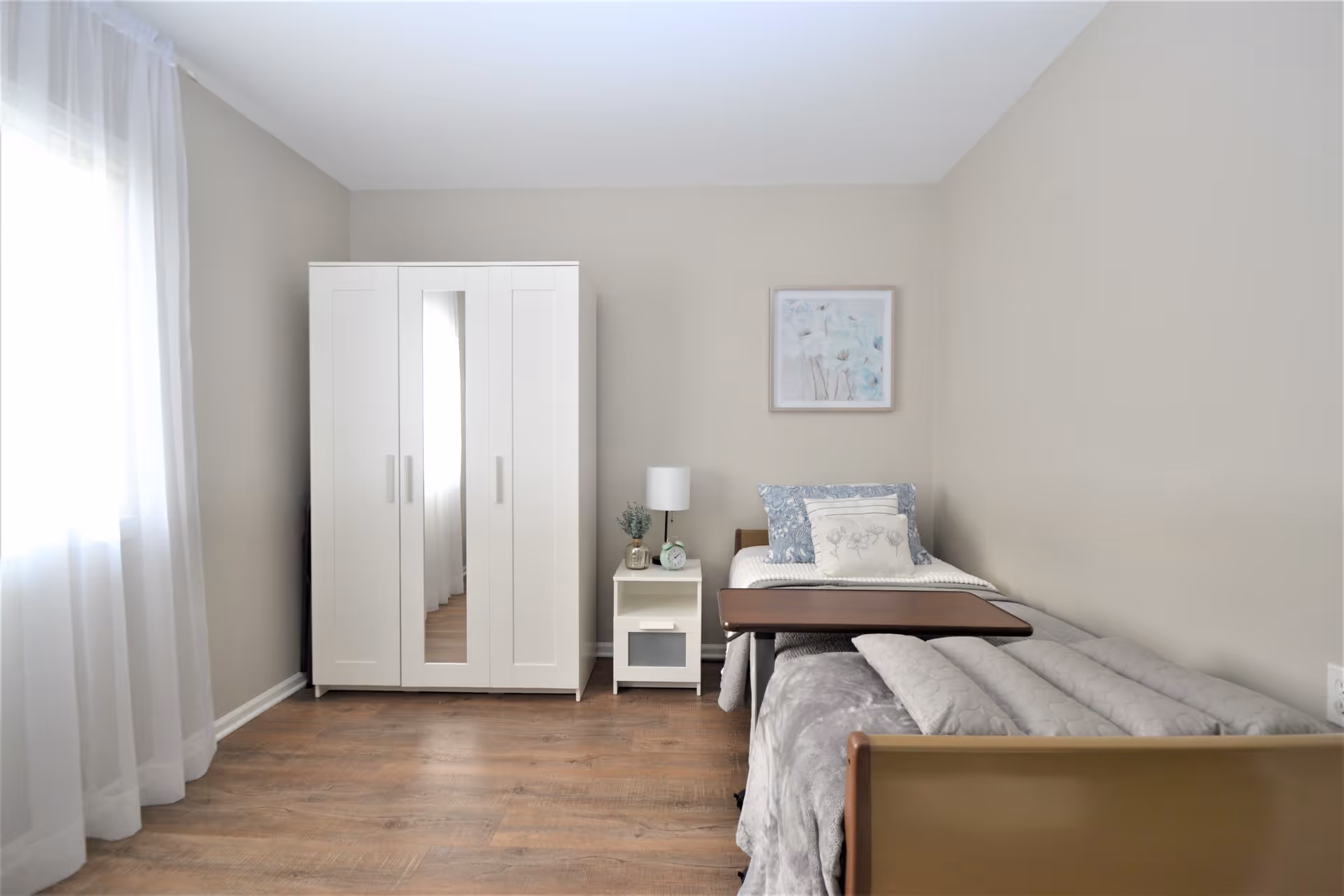 A simple, clean bedroom with a single bed covered in gray bedding and a tray table on top. Next to the bed is a white nightstand with a lamp, a small plant, and an alarm clock. A white wardrobe with a mirror in the center stands against the wall. Light beige walls and a window with sheer white curtains allow natural light to fill the room. A framed floral artwork hangs above the bed.