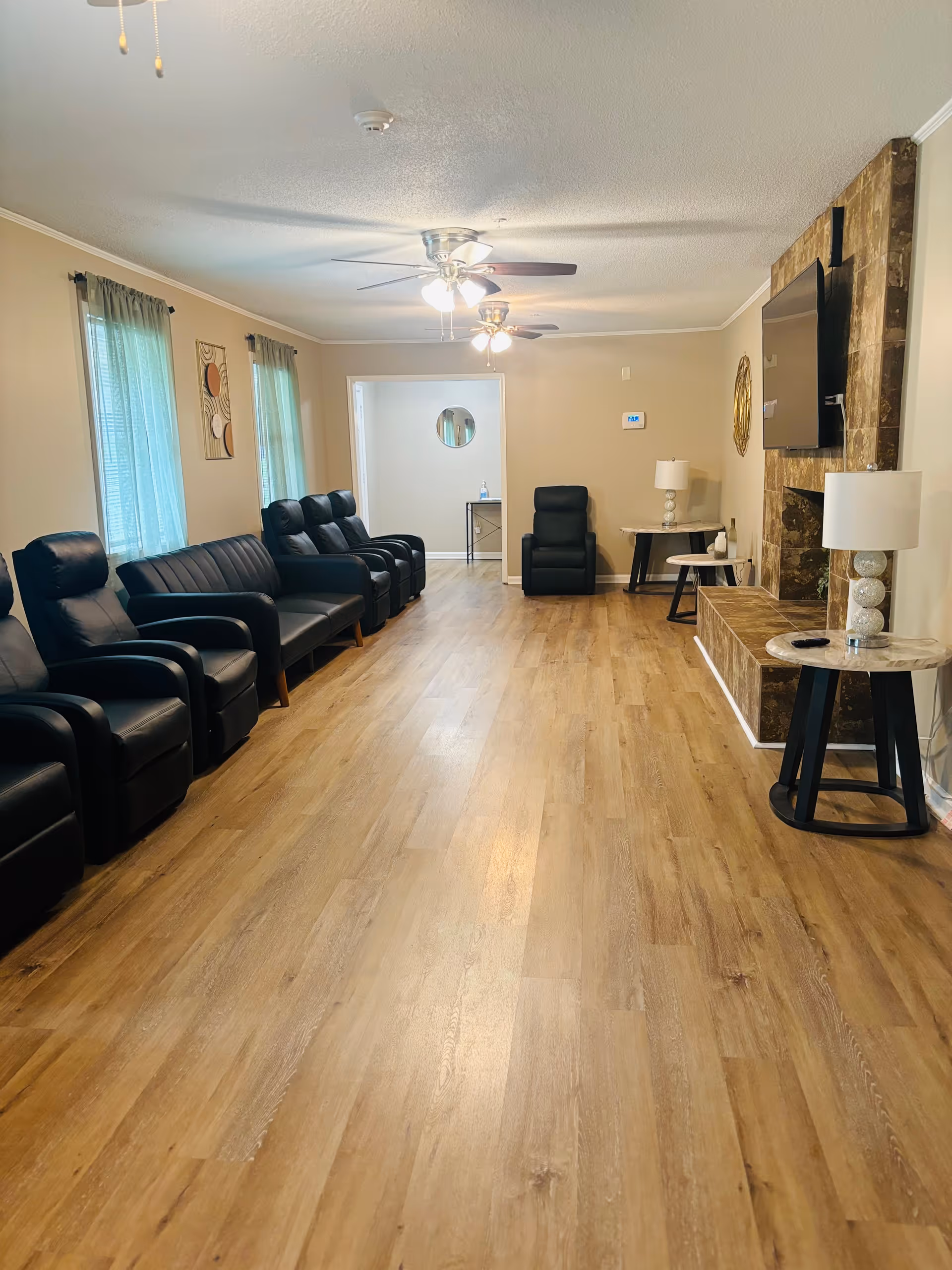 A spacious living room with wooden flooring, beige walls, and two ceiling fans with lights. The room features a row of black leather recliners and a black leather sofa along one wall with two windows covered by light blue curtains. On the opposite wall, there is a large flat-screen TV mounted above a stone fireplace. Two side tables with white lamps are placed near the fireplace. A small table with a mirror above it is visible in an adjacent room through an open doorway.