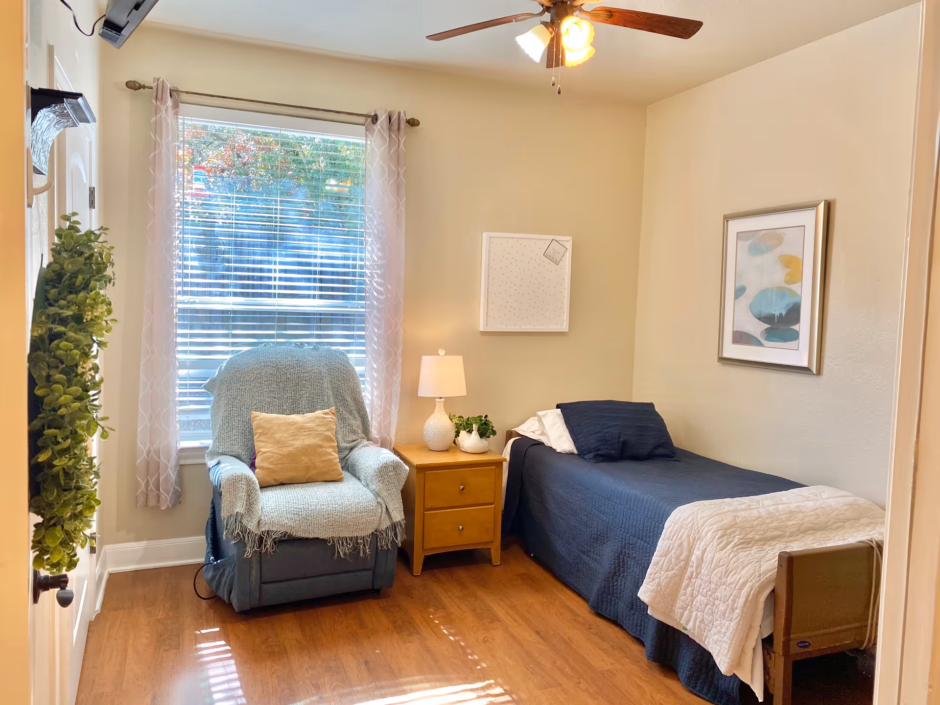 Bright assisted-living bedroom with a single bed, recliner by a window, a nightstand with lamp, and a ceiling fan.