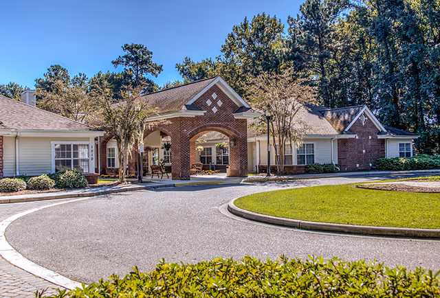 Front exterior view of a senior living facility named Oaks at Mt. Pleasant, featuring a circular driveway, brick and siding building with a covered entrance, benches, and trees under a clear blue sky.