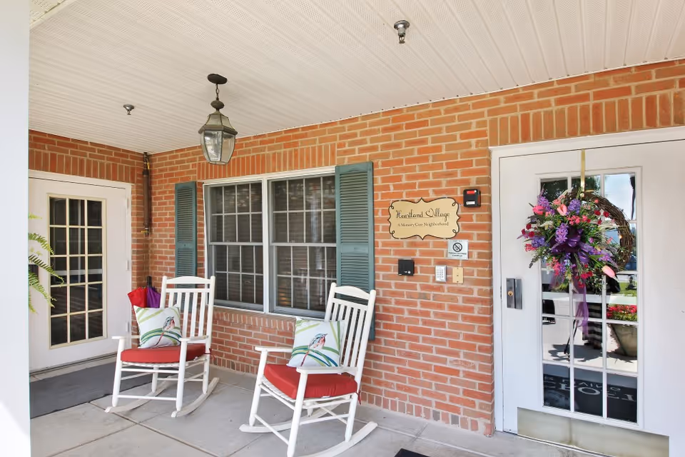 Covered outdoor porch area with two white rocking chairs with red cushions and bird-themed pillows, a brick wall with a window and green shutters, a hanging lantern light fixture, and two doors including one decorated with a floral wreath. A sign on the wall reads 'Heartland Village A Memory Care Neighborhood.'