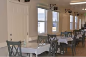 Interior view of a dining area in a retirement community with multiple tables covered with white tablecloths and green chairs. Large windows allow natural light to fill the room.