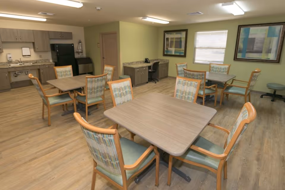 A dining area in a senior living facility featuring several square wooden tables surrounded by cushioned chairs with patterned backs. The room has light green walls, wood flooring, and two large framed abstract paintings. There is a kitchenette with cabinets, a sink, and a black refrigerator in the background, along with a small desk and office chair near a window with blinds.