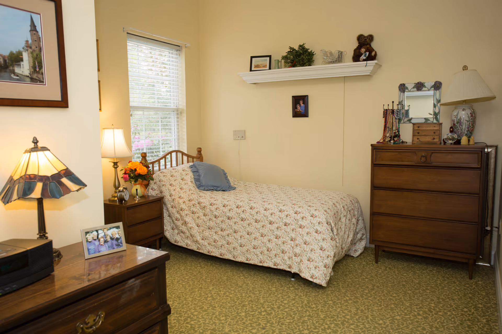A cozy bedroom with a single bed covered in a floral bedspread and a blue pillow. There is a wooden nightstand with a lamp and a vase of flowers next to the bed. A window with white blinds lets in natural light. On the right side, there is a wooden dresser with a lamp, a mirror, jewelry holders, and decorative items on top. The walls are light yellow, and there is a small shelf with plants and a teddy bear above the bed. A framed picture hangs on the wall to the left.