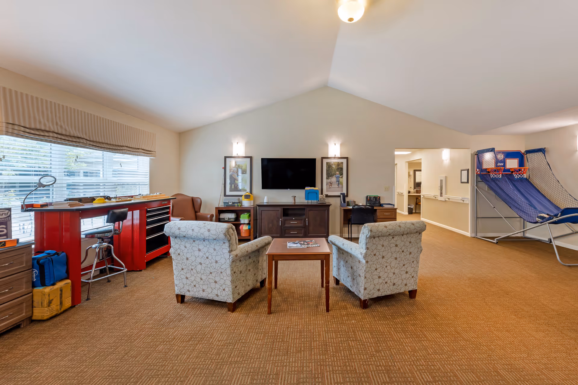 A spacious common area with two patterned armchairs facing a wooden coffee table and a TV mounted on the wall. The room has a high vaulted ceiling and beige carpet. To the left, there is a large window with blinds, a red workbench with a stool, and a brown armchair. To the right, there is a basketball arcade game. The walls are light-colored with two framed pictures flanking the TV.