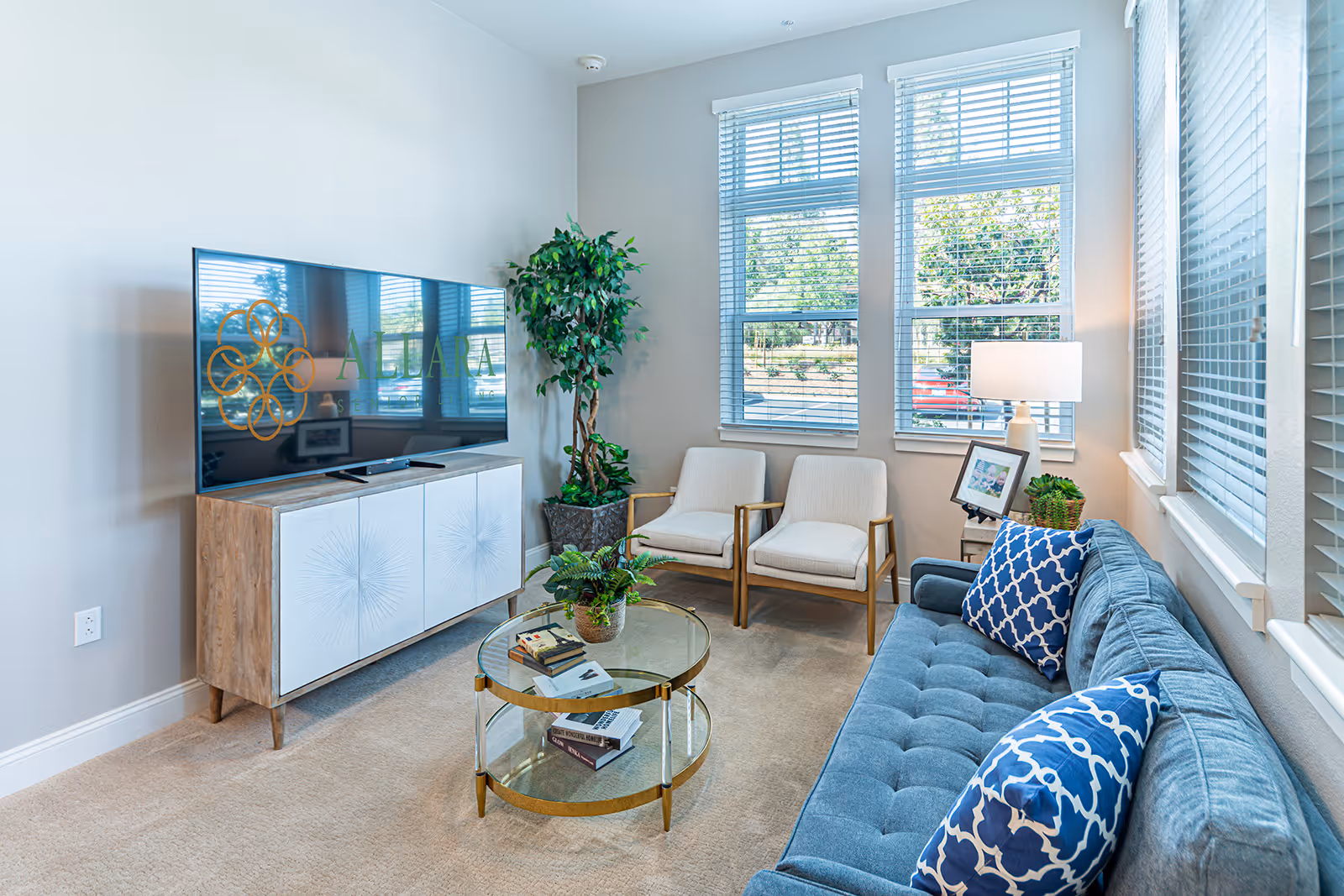 A bright and cozy living room with a blue tufted sofa adorned with two blue and white patterned pillows, a round glass coffee table with books and a plant, two beige armchairs with wooden arms, a wooden TV stand with a large flat-screen TV, a tall potted plant, and windows with white blinds letting in natural light.