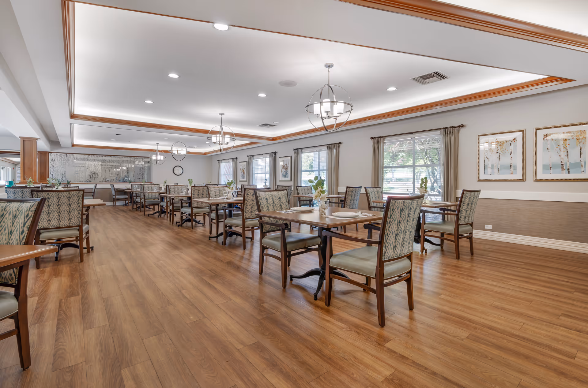 Spacious, well-lit dining room with multiple wooden tables and upholstered chairs on wood floors, pendant lighting, and windows with curtains.