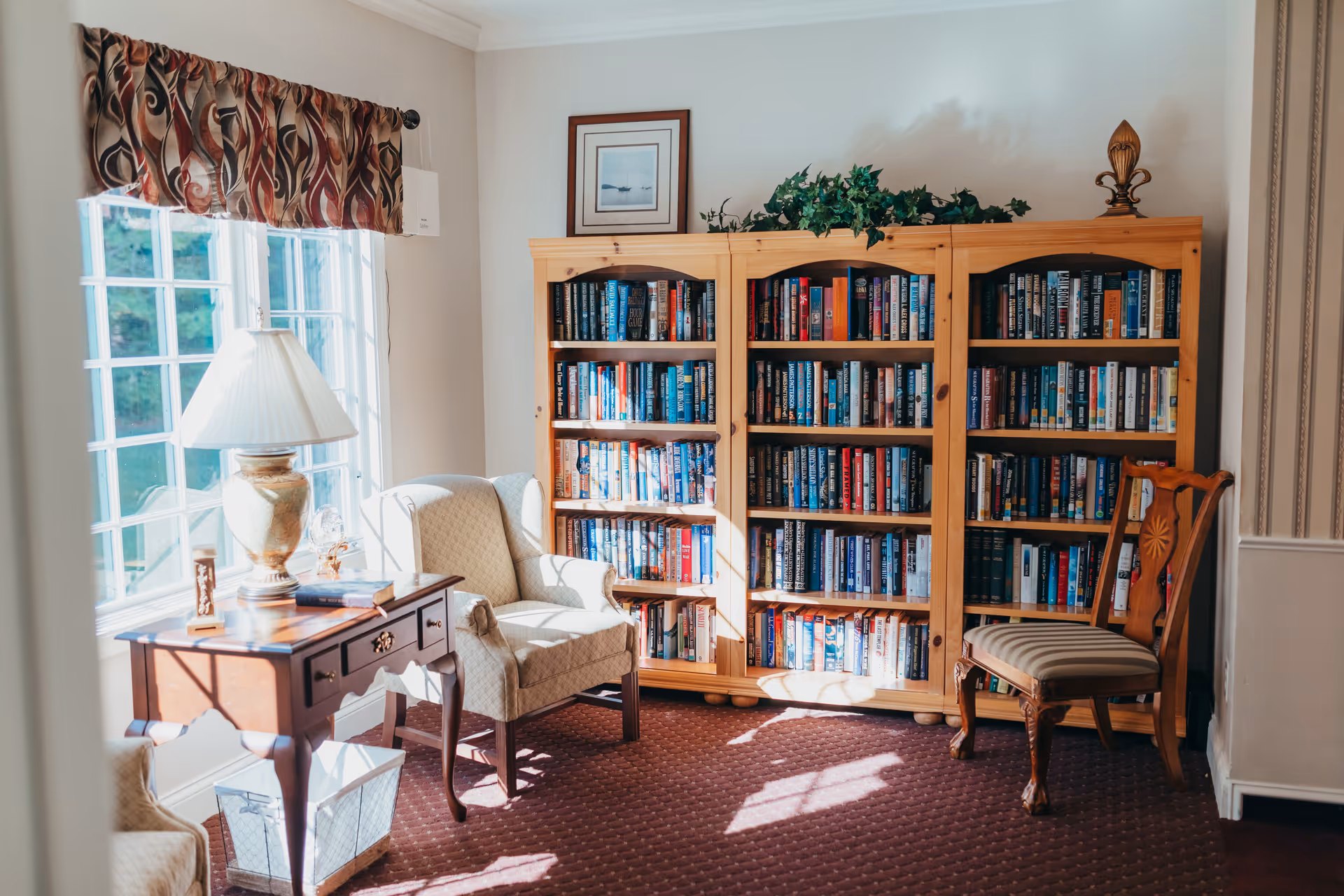 Sunlit reading room with armchairs, a wooden bookcase filled with books, and a side table with a lamp.