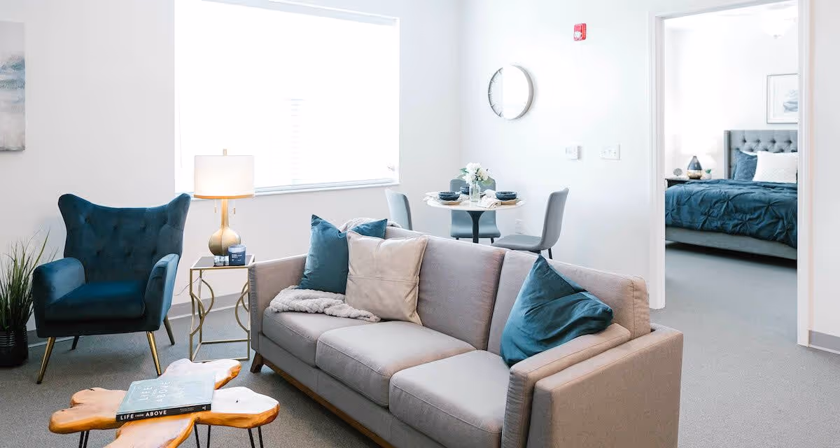 A bright living room with a gray sofa adorned with blue and beige pillows, a blue armchair, a wooden coffee table with a book on it, and a side table with a lamp. In the background, there is a small dining table set for two and a doorway leading to a bedroom with a blue bedspread.