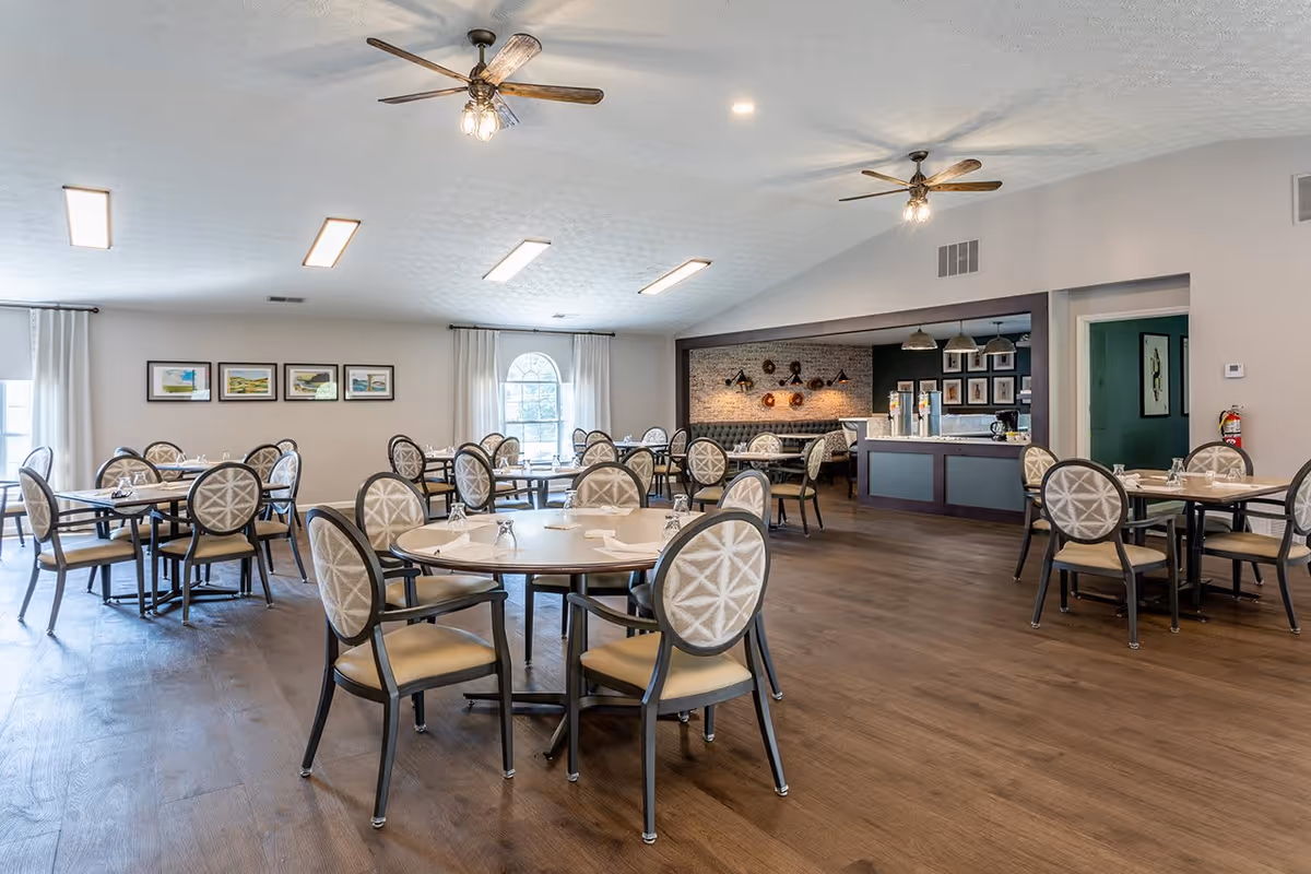 A spacious dining room with multiple round and rectangular tables surrounded by cushioned chairs with patterned backs. The room has wooden flooring, white walls, and several windows with white curtains letting in natural light. Ceiling fans and recessed lighting fixtures are visible on the ceiling. In the background, there is a counter area with beverage dispensers and decorative wall art.