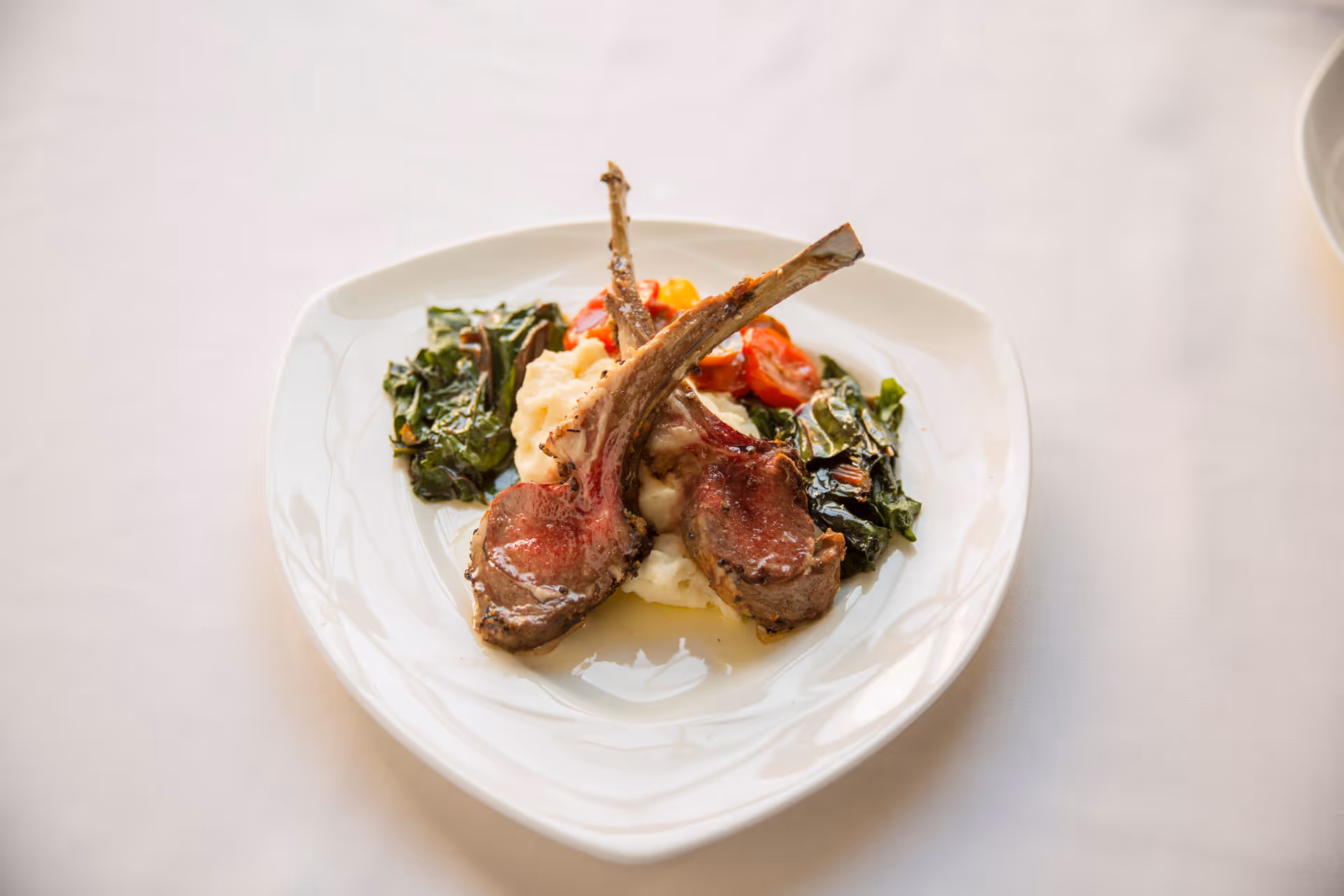 A white plate with two cooked lamb chops served on a bed of mashed potatoes, accompanied by sautéed greens and roasted cherry tomatoes, placed on a white tablecloth.