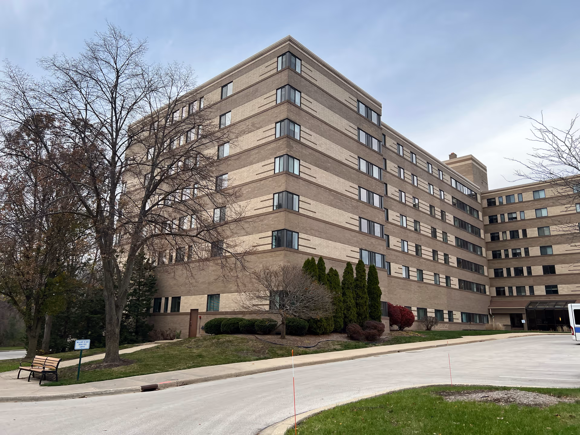 Exterior view of a multi-story senior living facility building with beige and brown brickwork. The building has many windows and is surrounded by trees, shrubs, and a small grassy area. A bench and a sign are visible near the sidewalk, and a driveway curves around the front of the building under a cloudy sky.