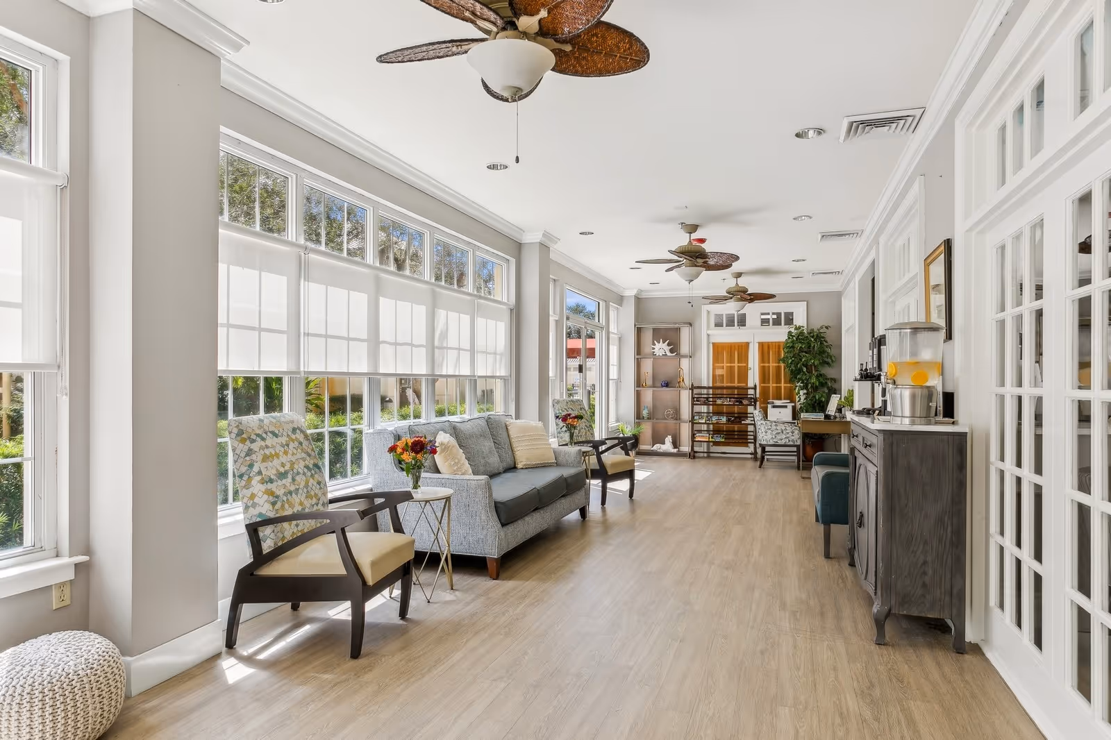 A bright and spacious living room area with large windows allowing natural light to fill the space. The room features a gray sofa with cushions, two armchairs, a small side table with a vase of flowers, ceiling fans with leaf-shaped blades, a wooden cabinet with a beverage dispenser, and decorative shelves with plants and ornaments. The floor is light wood, and the walls are painted white.