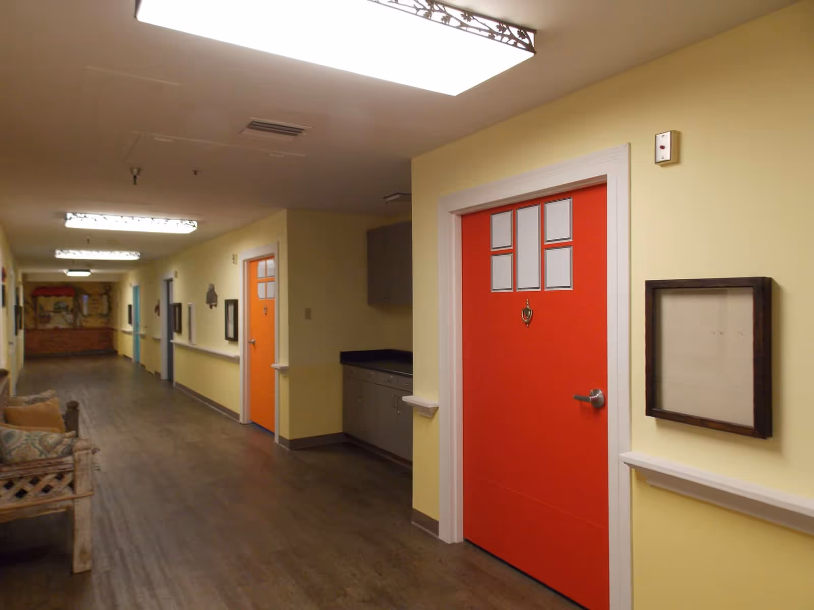 A long, well-lit interior hallway with yellow walls, wood flooring, seating, and red doors.
