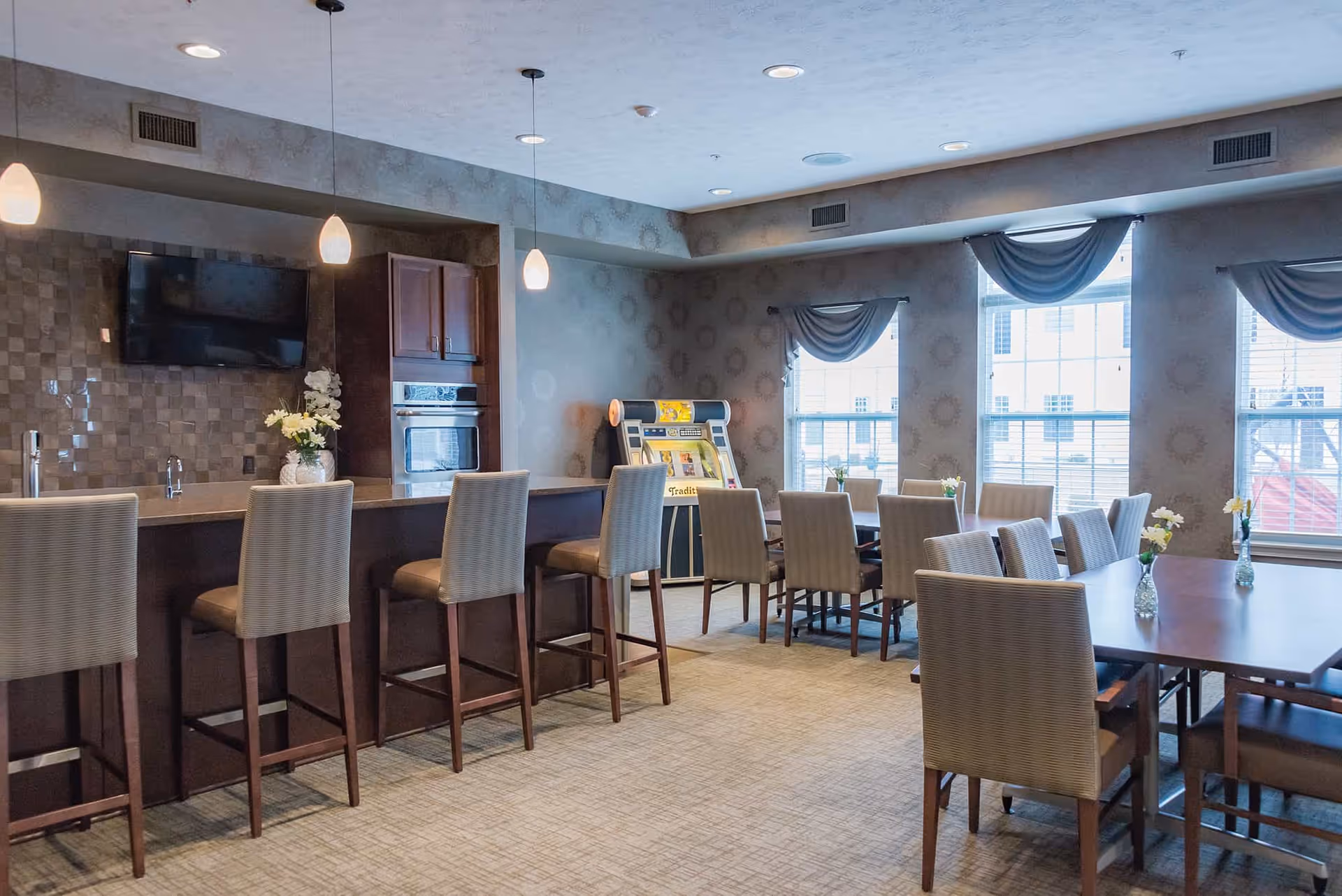 Communal dining and lounge area with a long bar, high stools, dining tables and chairs, windows, and a jukebox.