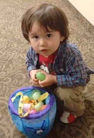 A young child with brown hair wearing a plaid shirt, red t-shirt, and beige pants is crouching on a carpeted floor holding a green plastic egg. In front of the child is a blue basket filled with colorful plastic eggs.
