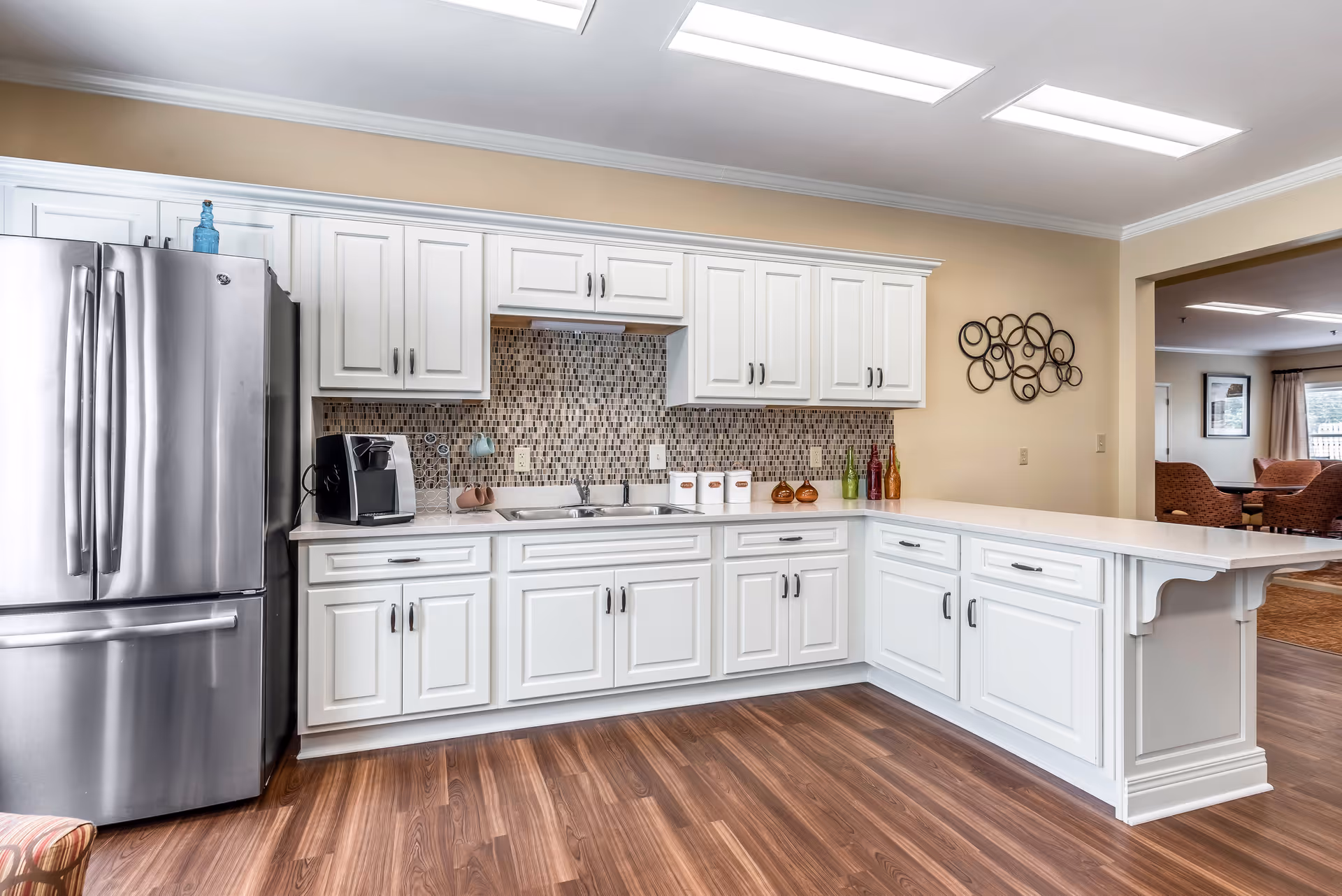 A modern kitchen area with white cabinetry, a stainless steel refrigerator, a coffee maker on the counter, a mosaic tile backsplash, and a white countertop extending into a breakfast bar. The kitchen has wood flooring and is brightly lit with ceiling lights. In the background, there is a glimpse of a living area with chairs and framed pictures on the wall.