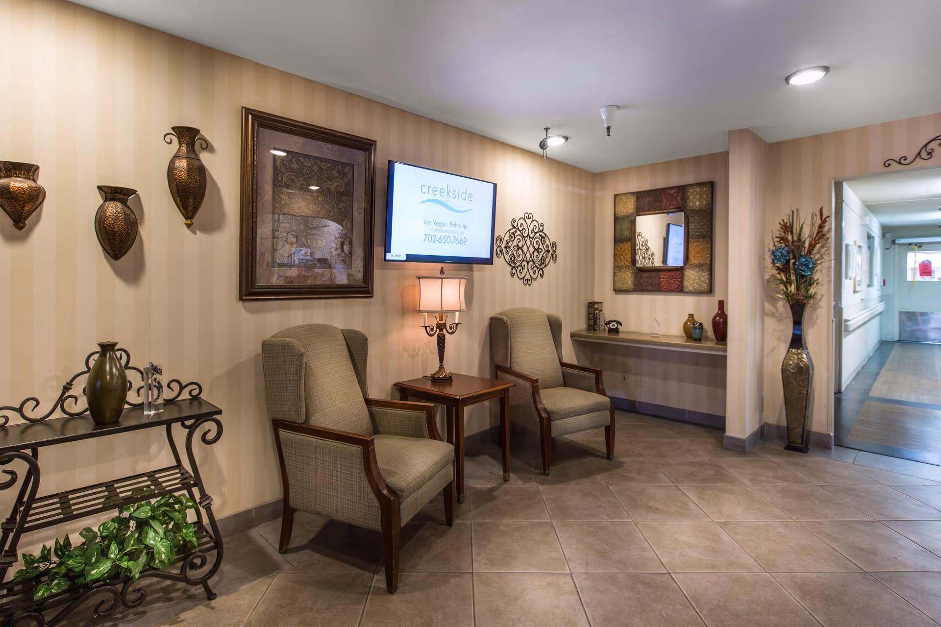 A cozy seating area in a hallway with two upholstered armchairs separated by a wooden side table with a lamp. The walls have beige vertical stripes and are decorated with framed artwork, a decorative metal wall piece, and a large mirror. A small black metal table with decorative vases and a green plant is on the left. A tall vase with flowers is on the right near the hallway entrance.