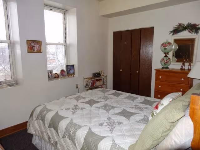 A bedroom with a bed covered in a white and gray patterned quilt, green pillows, and a wooden headboard. There are two windows with blinds on the left wall, a small shelf with framed photos and books beneath the windows, and a wooden dresser with a decorative lamp and mirror on the right side. A closed wooden closet door is visible in the background.