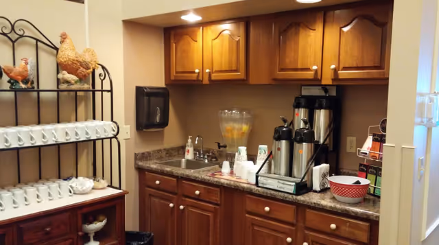 Small kitchenette-style coffee station with wooden cabinets, countertop coffee urns, a pitcher of infused water, and shelves of white mugs.