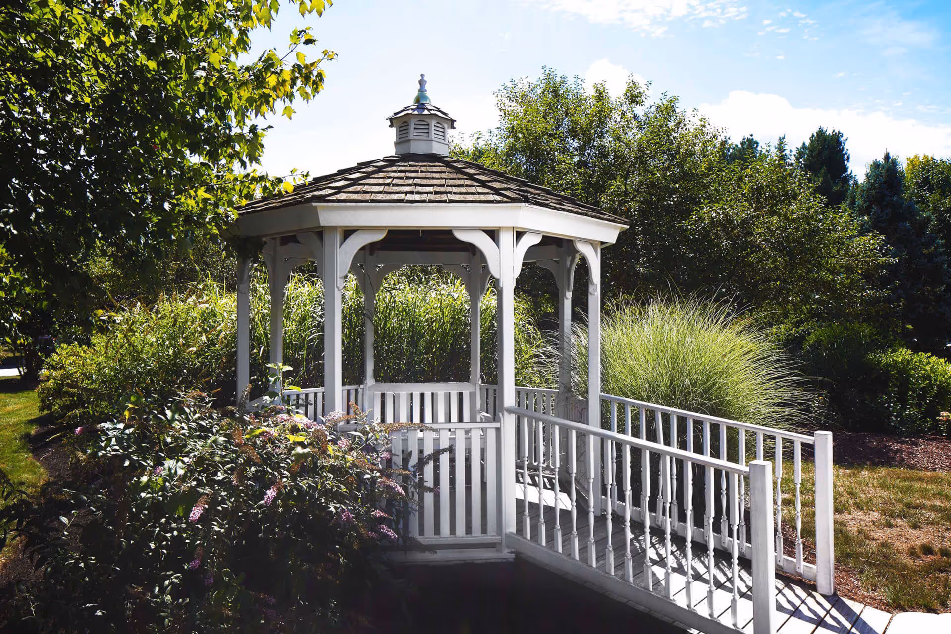 A white wooden gazebo with a shingled roof surrounded by lush greenery and bushes under a partly cloudy sky. There is a white railing leading up to the gazebo, which is situated in a garden area.