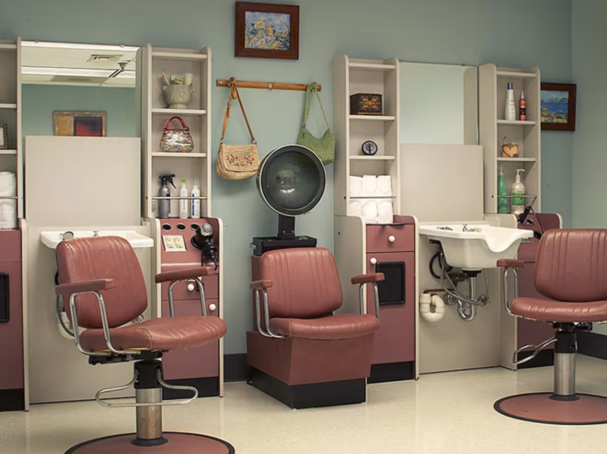 Interior view of a salon area with three vintage-style maroon salon chairs in front of mirrors and shelves. The shelves hold various hair care products, towels, and decorative items. A hair dryer hood is positioned between two of the chairs, and a sink for washing hair is visible on the right side.