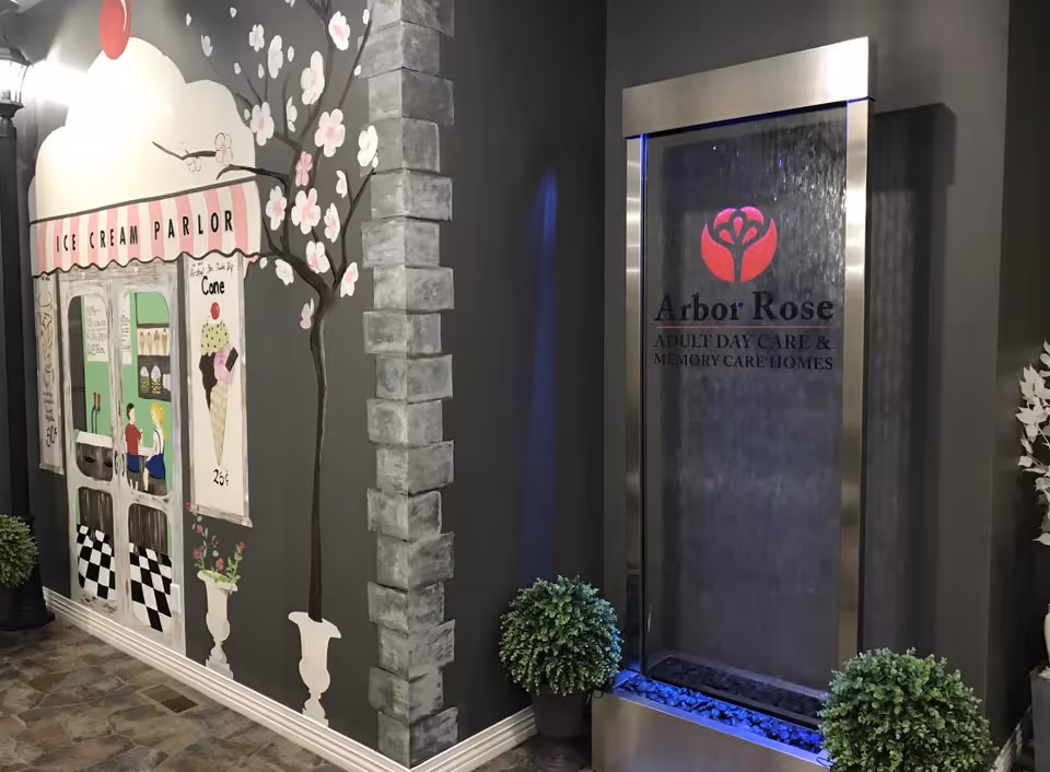 Interior view of a senior living facility featuring a decorative wall mural of an ice cream parlor with a cherry on top, a flowering tree, and a faux brick column. Next to the mural is a modern water feature with the Arbor Rose logo and the text 'Adult Day Care & Memory Care Homes'. Two potted plants are placed near the water feature.