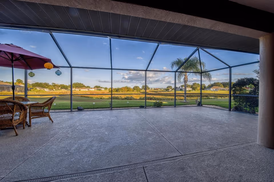 A spacious screened-in patio area with a concrete floor, featuring a wicker table and chairs with a red umbrella and hanging lanterns, overlooking a green lawn, palm trees, and a scenic landscape under a blue sky with some clouds.