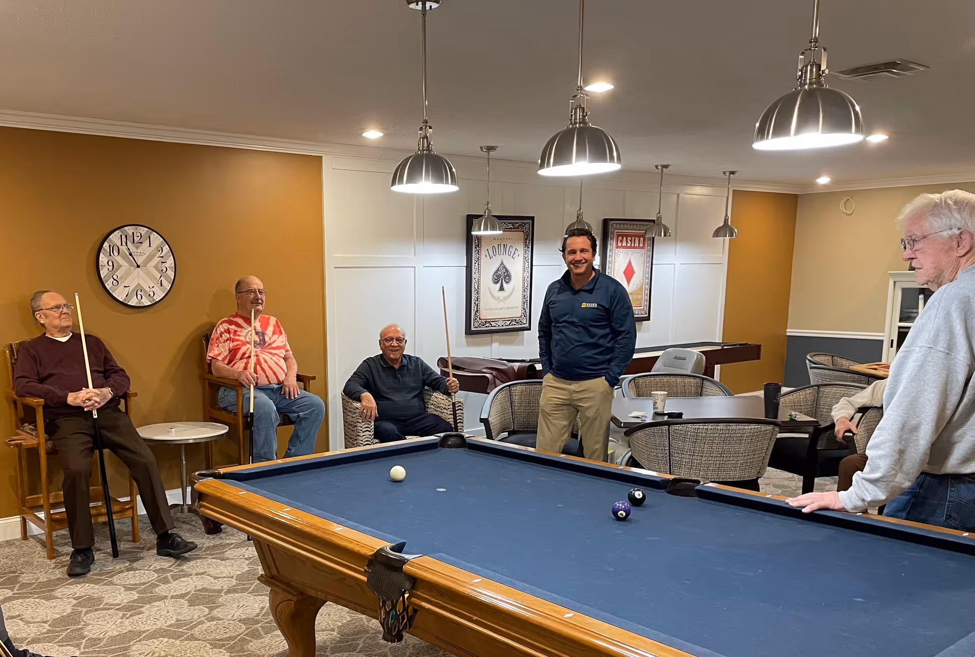 Several men standing and sitting around a pool table in a well-lit senior living facility game room.
