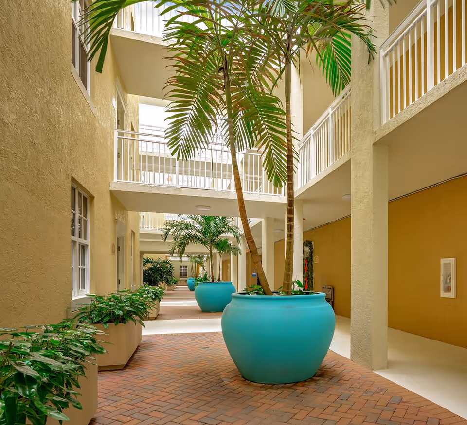 Indoor corridor area of a senior living facility with large turquoise planters containing tall palm trees and smaller plants in rectangular beige planters along the walls. The corridor has a brick floor and beige walls with white railings on the upper floors.