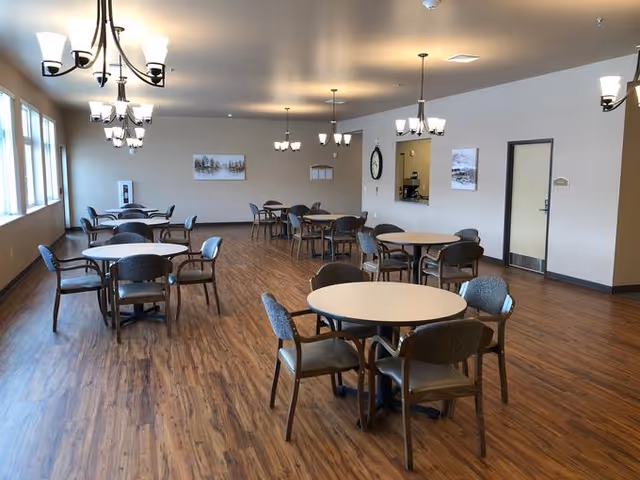 A spacious dining room with multiple round tables and chairs arranged neatly on a wooden floor. The room is well-lit with large windows on one side and several ceiling light fixtures. There are two framed pictures on the far wall, a clock, and a door on the right side.