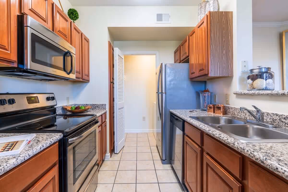 A modern kitchen with wooden cabinets, granite countertops, a stainless steel microwave, stove, refrigerator, and dishwasher. The kitchen has a double sink, tiled floor, and decorative items on the counter.