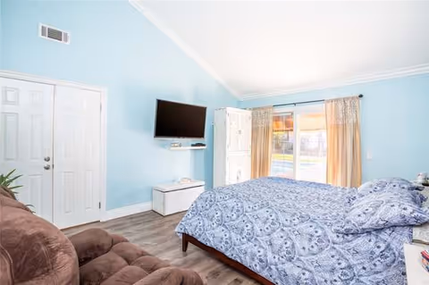 A bright bedroom with light blue walls, a large bed with blue patterned bedding, a brown recliner chair, a wall-mounted flat screen TV, a white door, and a sliding glass door with beige curtains letting in natural light.