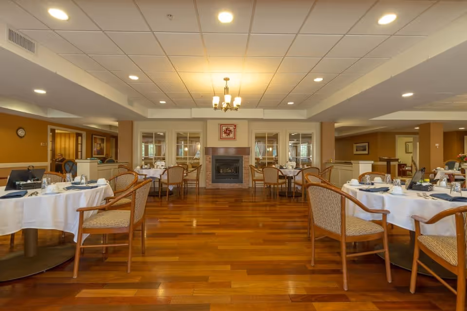 A spacious dining room with wooden floors and round tables covered with white tablecloths. Each table is set with plates, cups, and silverware, and surrounded by wooden chairs with cushioned seats. A fireplace is centered on the far wall, flanked by windows with white curtains. The ceiling has recessed lighting and a chandelier above the fireplace.